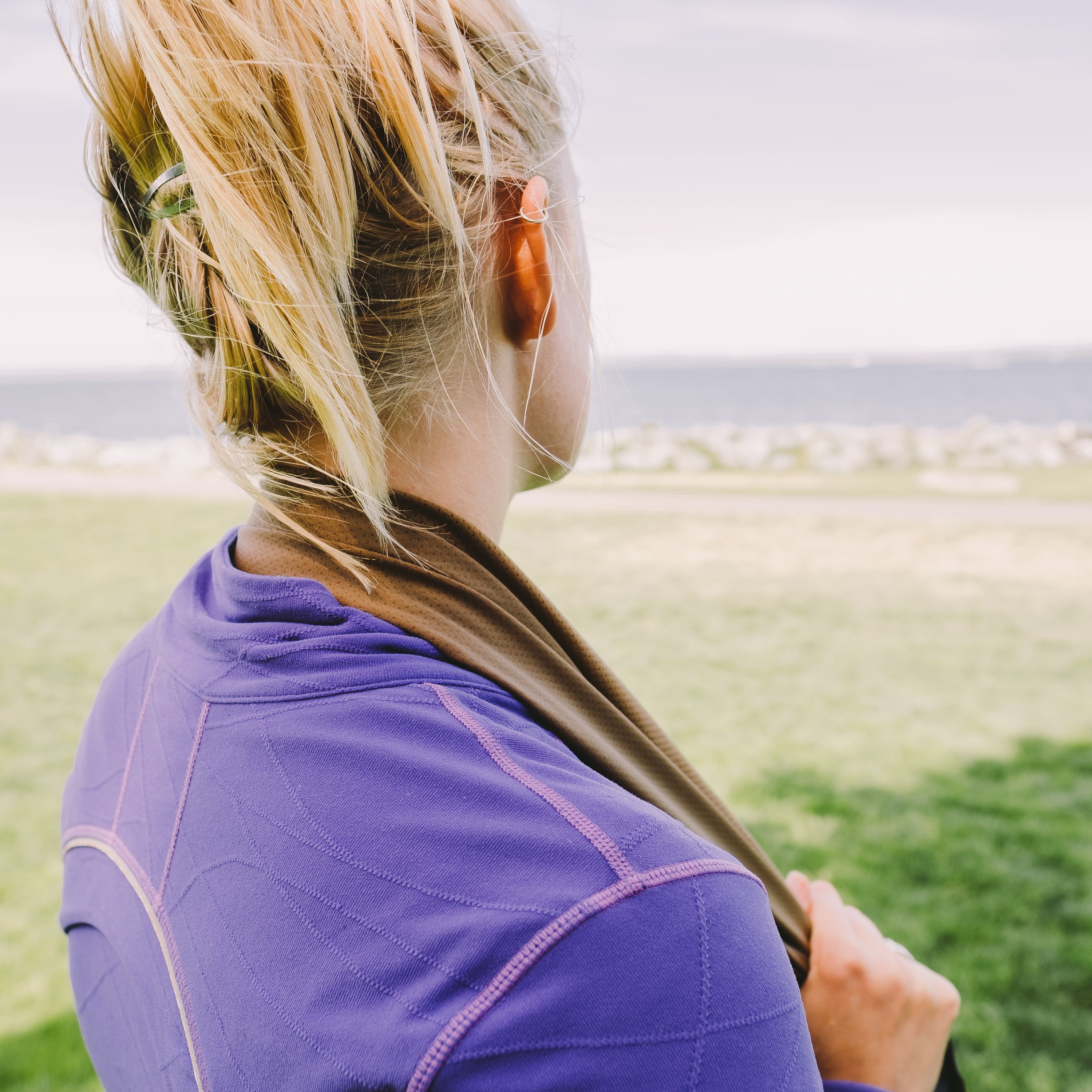 Woman facing a bay of water wearing a purple shirt with a cooling towel wrapped around her neck