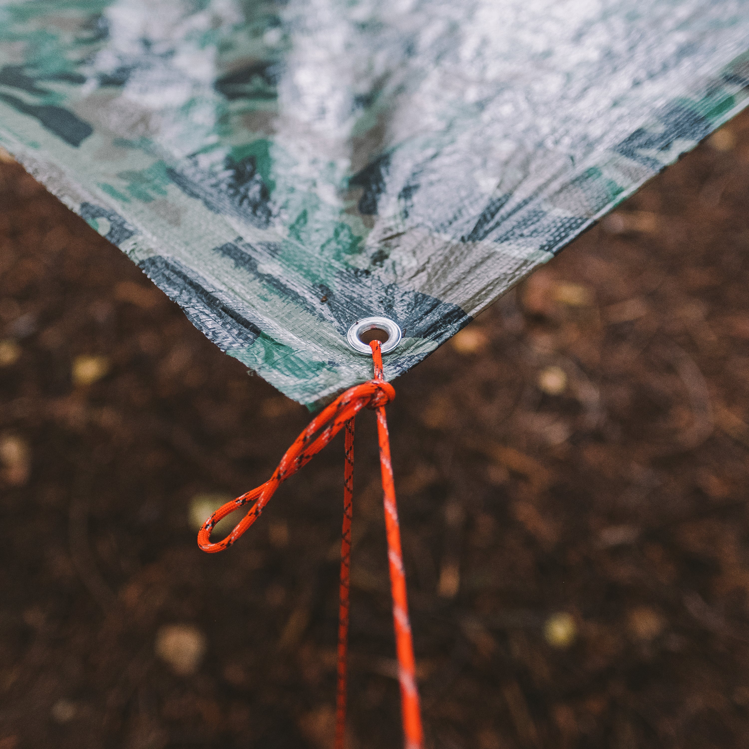 A piece of orange paracord attached to a corner of a camoflauge tarp and pulling it taut, brown dirt/ground in the background.