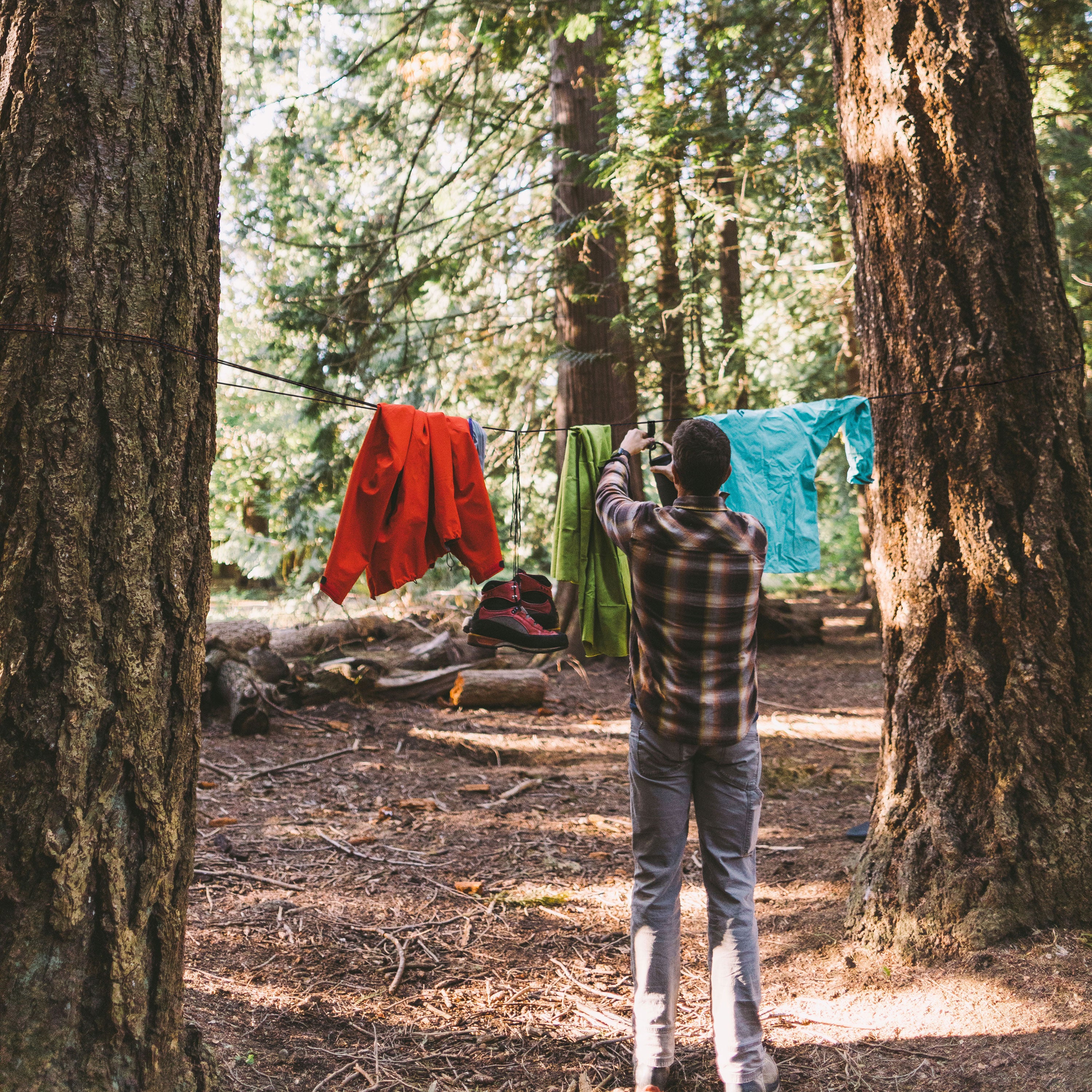 A man hanging a water bottle up on a clothesline made of GEAR AID paracord against two trees in the woods.