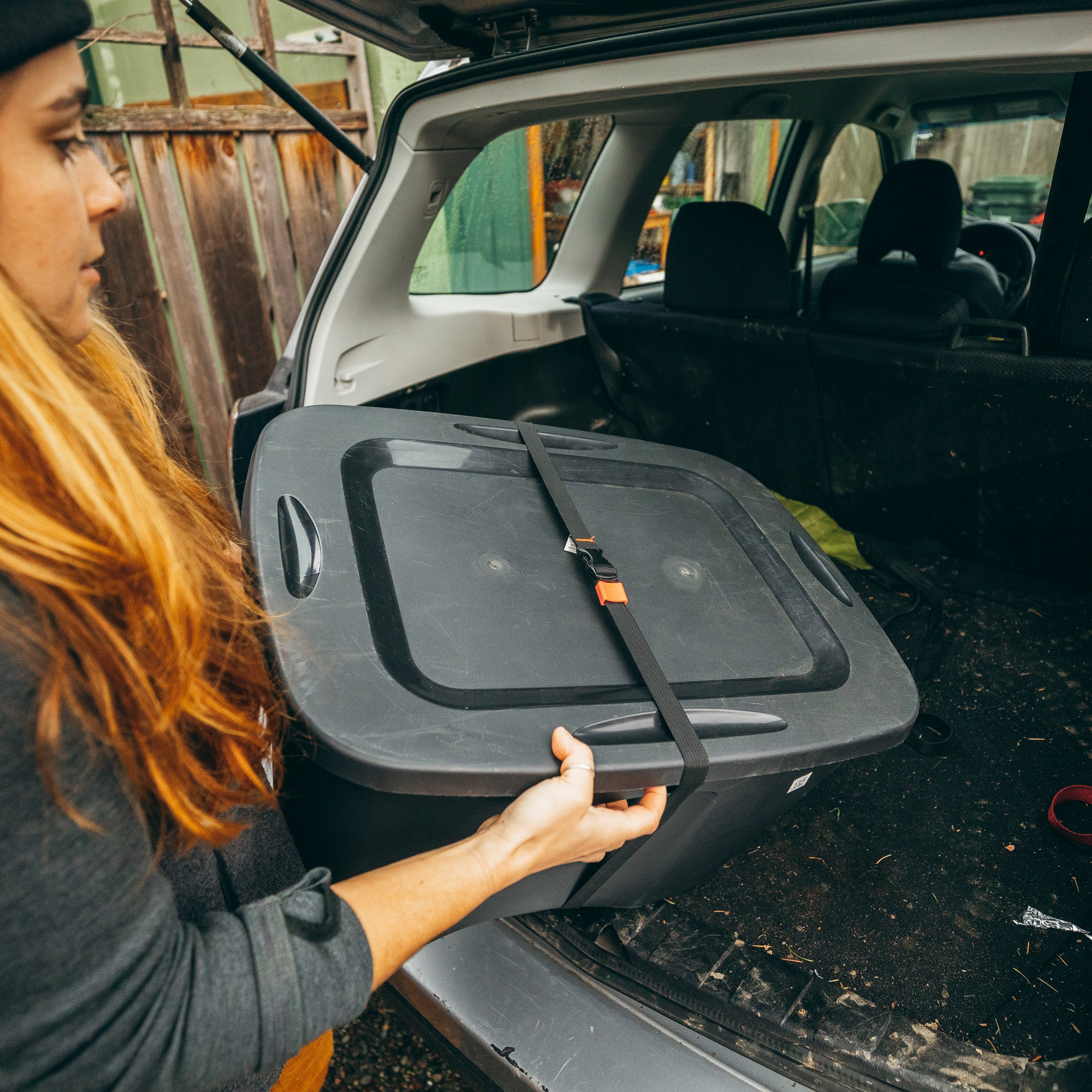 Person putting a storage tote wrapped in a Utility Strap into the back of an SUV, fence in background