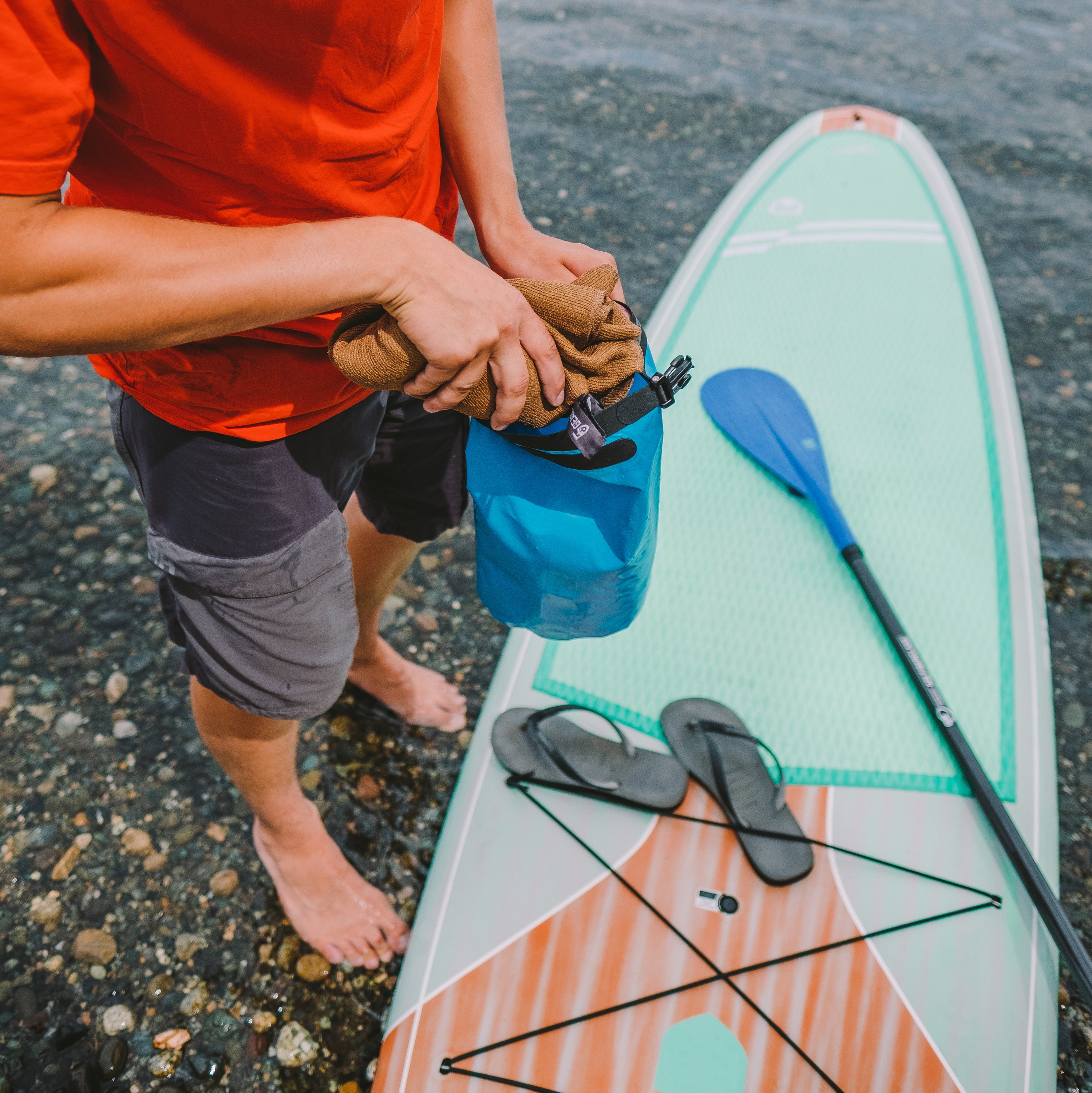 Person stuffing a rolled up coyote colored micro terry towel into a blue dry bag, paddleboard and flip flops are beneath them on a rocky beach by the water.