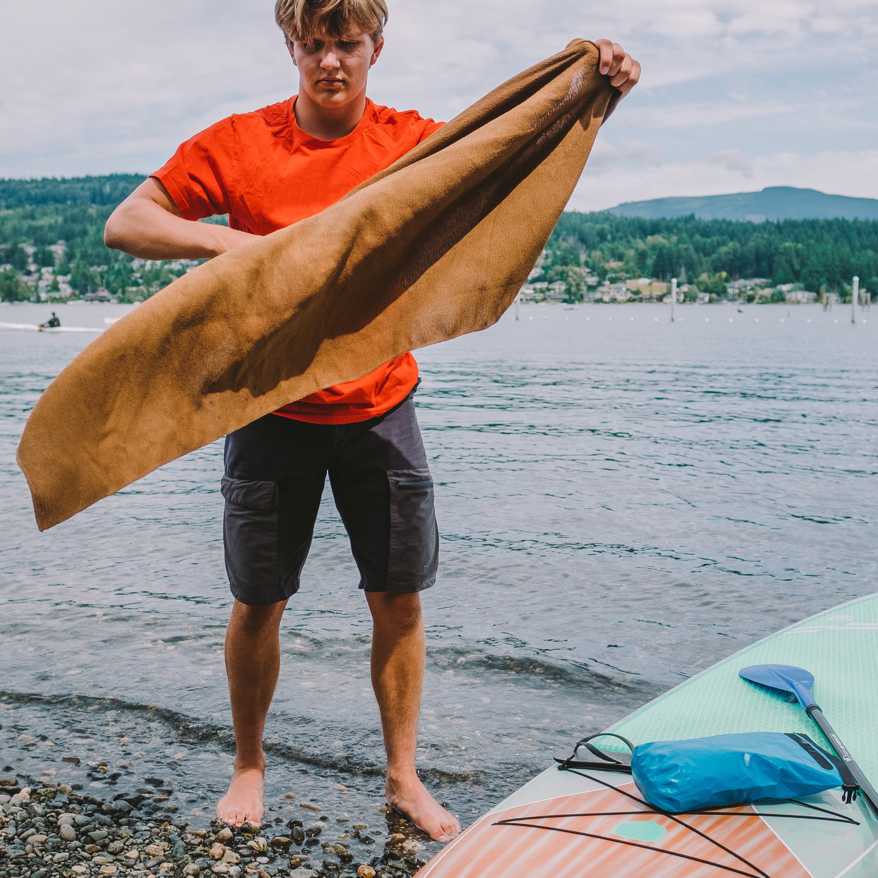 Man at a rocky beach with green hills in the background next to a paddleboard and dry bag displaying a coyote microterry towel that is blowing in the wind